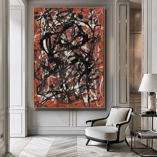 A large framed abstract canvas print with black and white chaotic brushstrokes over a terracotta orange-red background, displayed on a gray paneled wall in a modern living room setting with a white armchair and side table.