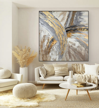 A gold and grey abstract painting displayed on a wall above a modern living room setup with neutral colored furniture and decorative items.