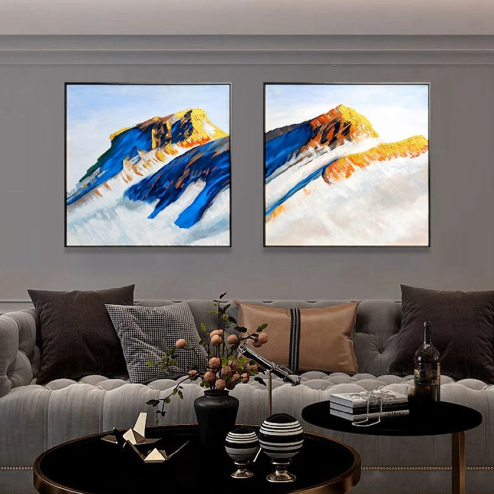 Room mockup showing two mountain paintings with deep blue slopes and ochre peaks rising above broad pale white areas.