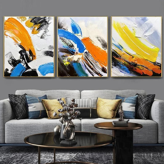 Room mockup showing three expressive abstracts with broad blue, orange and yellow strokes over white fields with black accents.