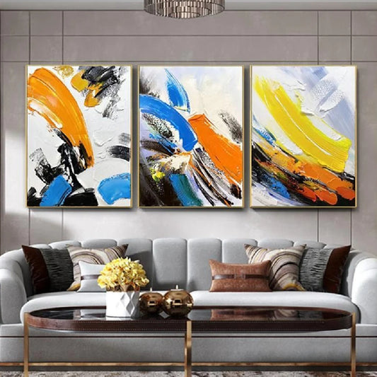Room mockup showing three expressive abstracts with broad blue, orange and yellow strokes over white fields with black accents.