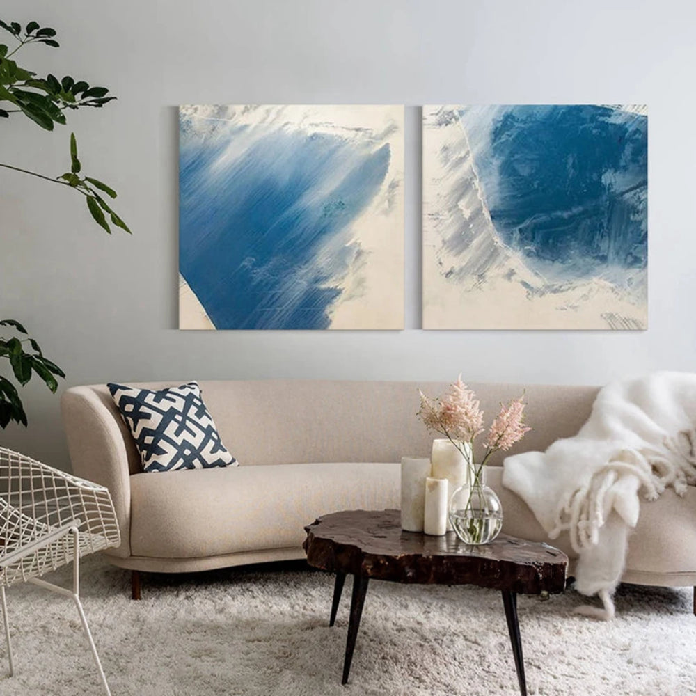 Room mockup showing two square abstract paintings with broad blue brushstrokes and pale feathered edges on off-white backgrounds.