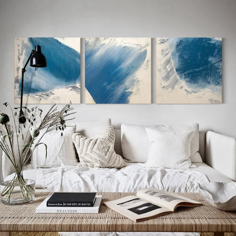 Room mockup showing three square abstracts with broad blue brushstrokes and feathered pale edges on off-white backgrounds.