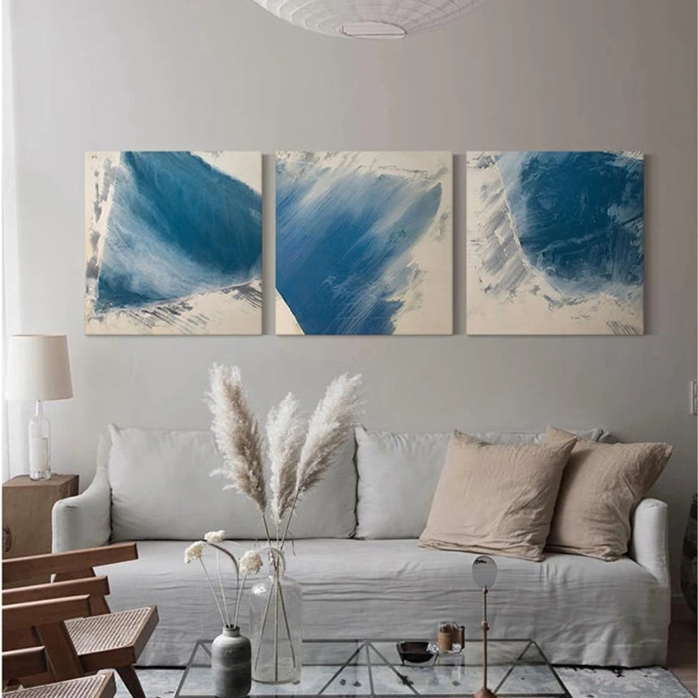 Room mockup showing three square abstracts with broad blue brushstrokes and feathered pale edges on off-white backgrounds.